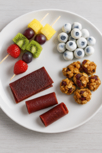 A white plate with four types of homemade fruit snacks including fruit kabobs, yogurt-covered blueberries, fruit leather, and oat-nut clusters.