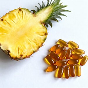 A close-up of amber-colored softgel pineapple enzyme supplement capsules next to a fresh, sliced pineapple on a clean, white background.